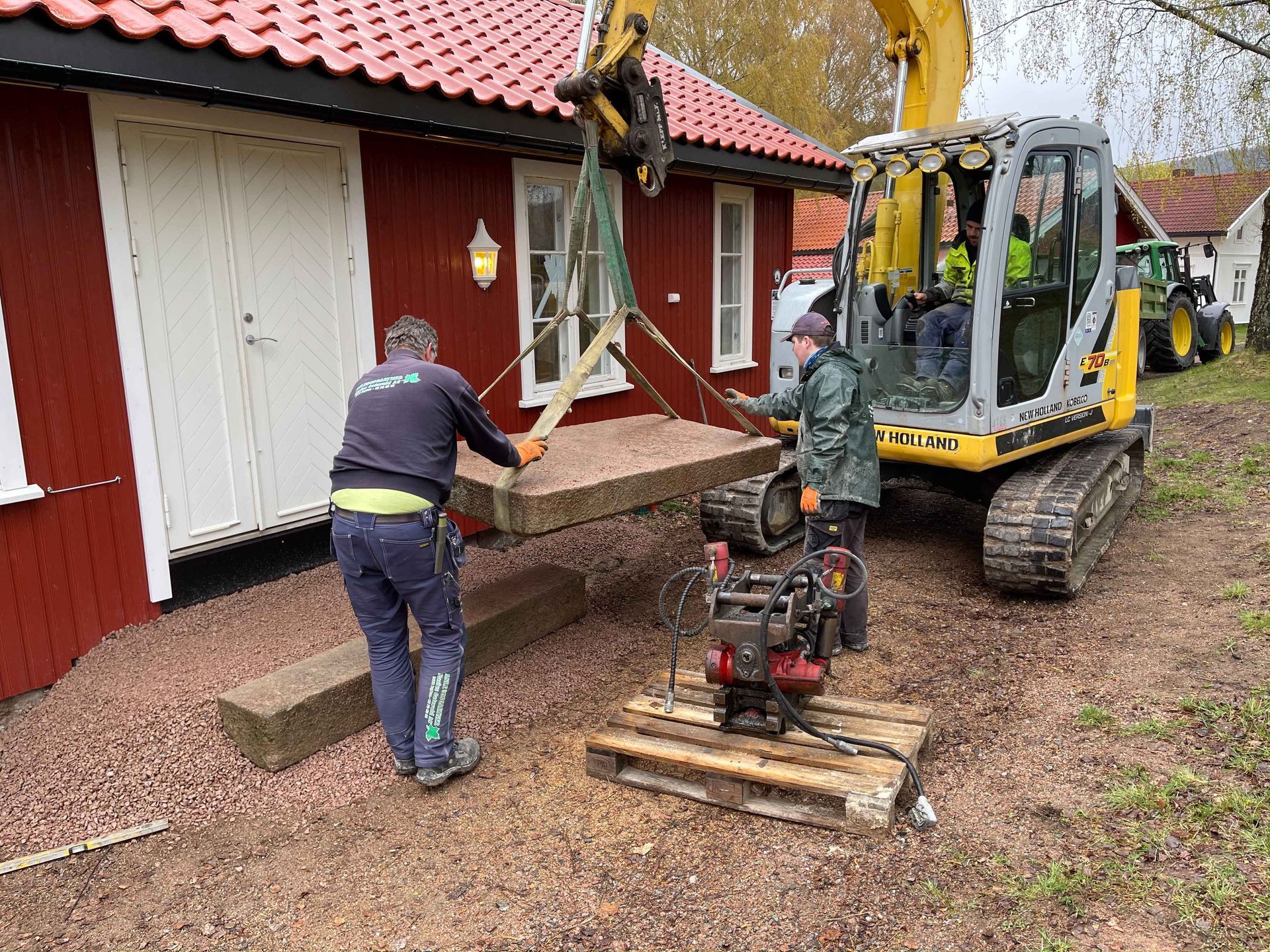 Arbeidere bruker en gravemaskin til å løfte en steinplate nær en rød bygning.