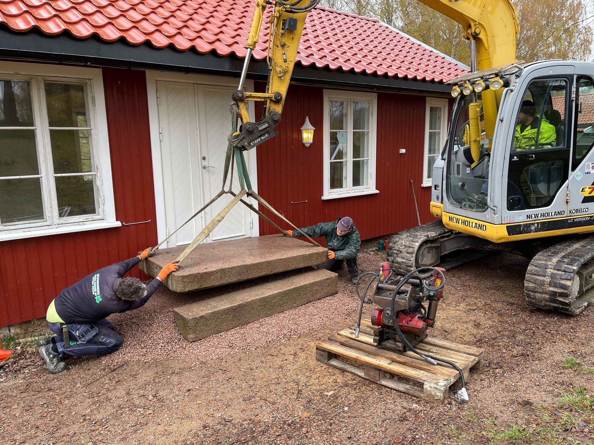Arbeidere bruker gravemaskin til å løfte steintrapper foran et rødt hus.