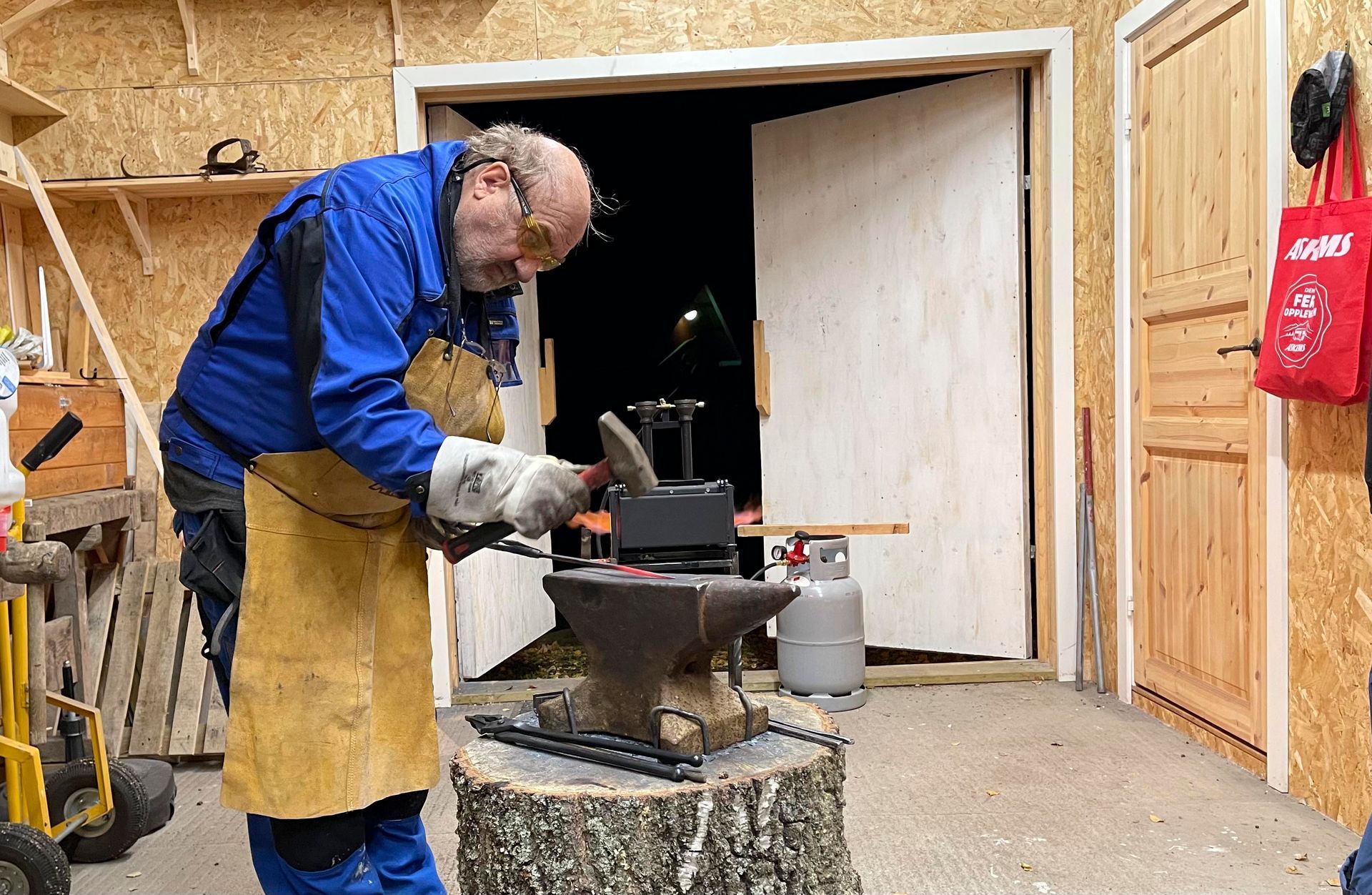 Smed i arbeid, smir metall på en ambolt, iført verneutstyr inne i et verksted.