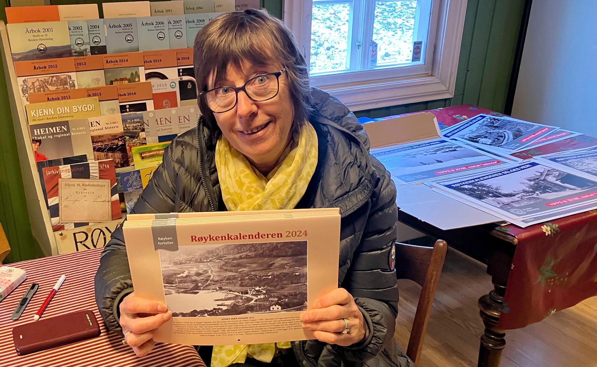 Kvinne som holder et gammelt fotoalbum, smilende. Bak henne, en skjerm med gamle bilder. Innendørs.