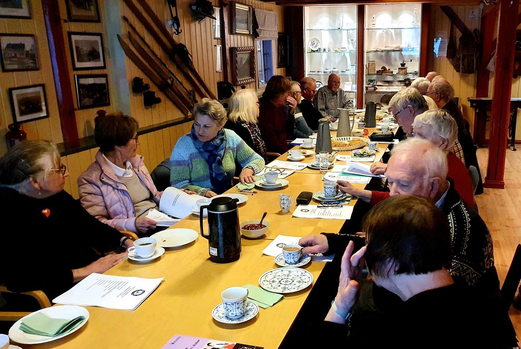 Gruppe mennesker ved et langt bord drikker kaffe, inne i et rom med trepanel.