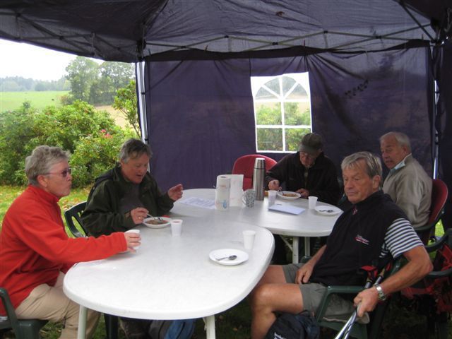 En gruppe mennesker sitter rundt et bord i et telt utendørs, spiser og snakker.