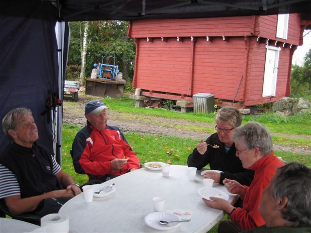 Gruppe mennesker sitter ved et bord og spiser utendørs, med en rød bygning i bakgrunnen.