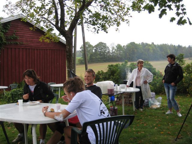 Folk spiser ved hvite bord utendørs, rød låve i bakgrunnen.
