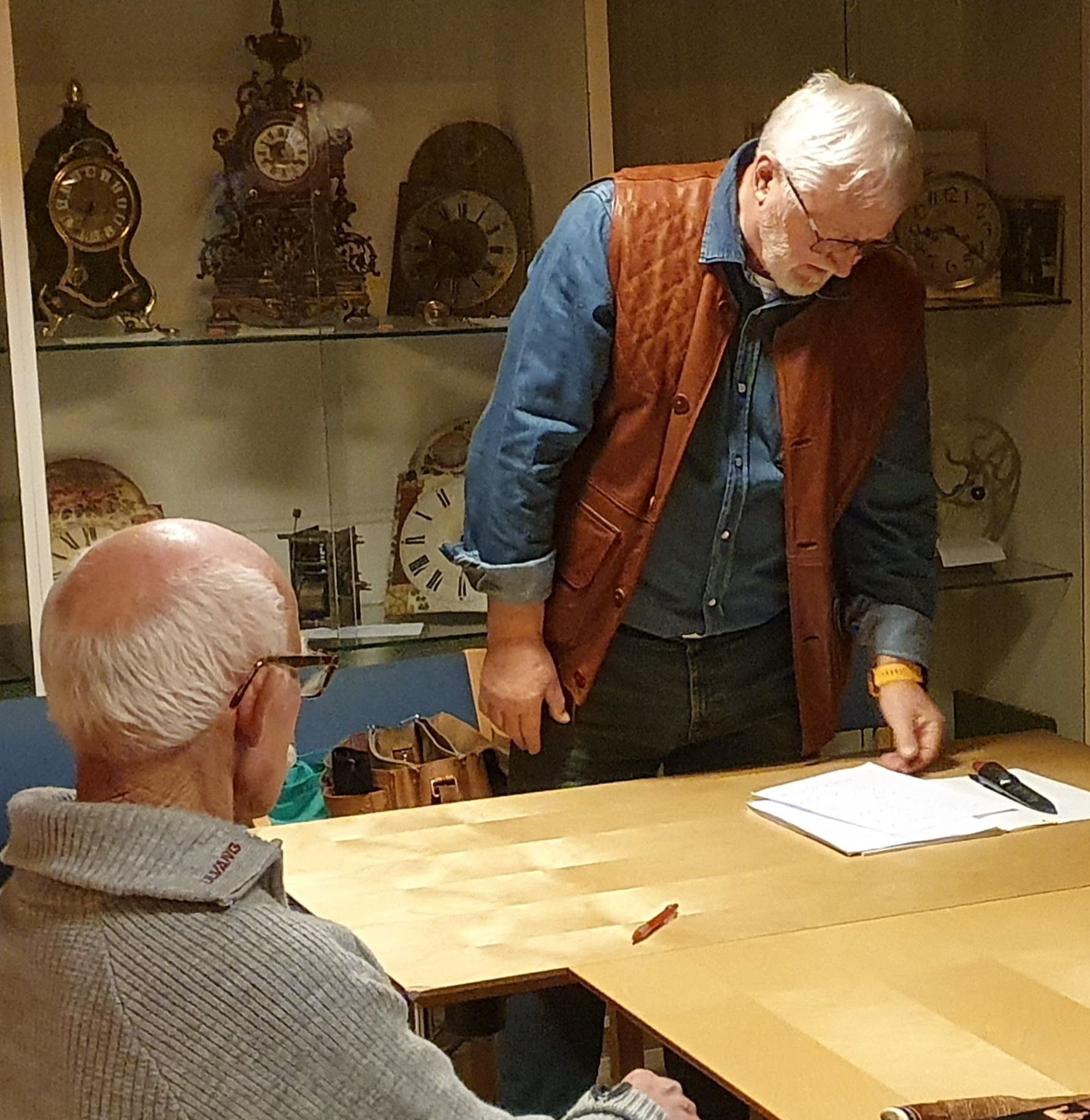 To menn undersøker papirer ved et bord. Klokker står utstilt i et skap bak dem.