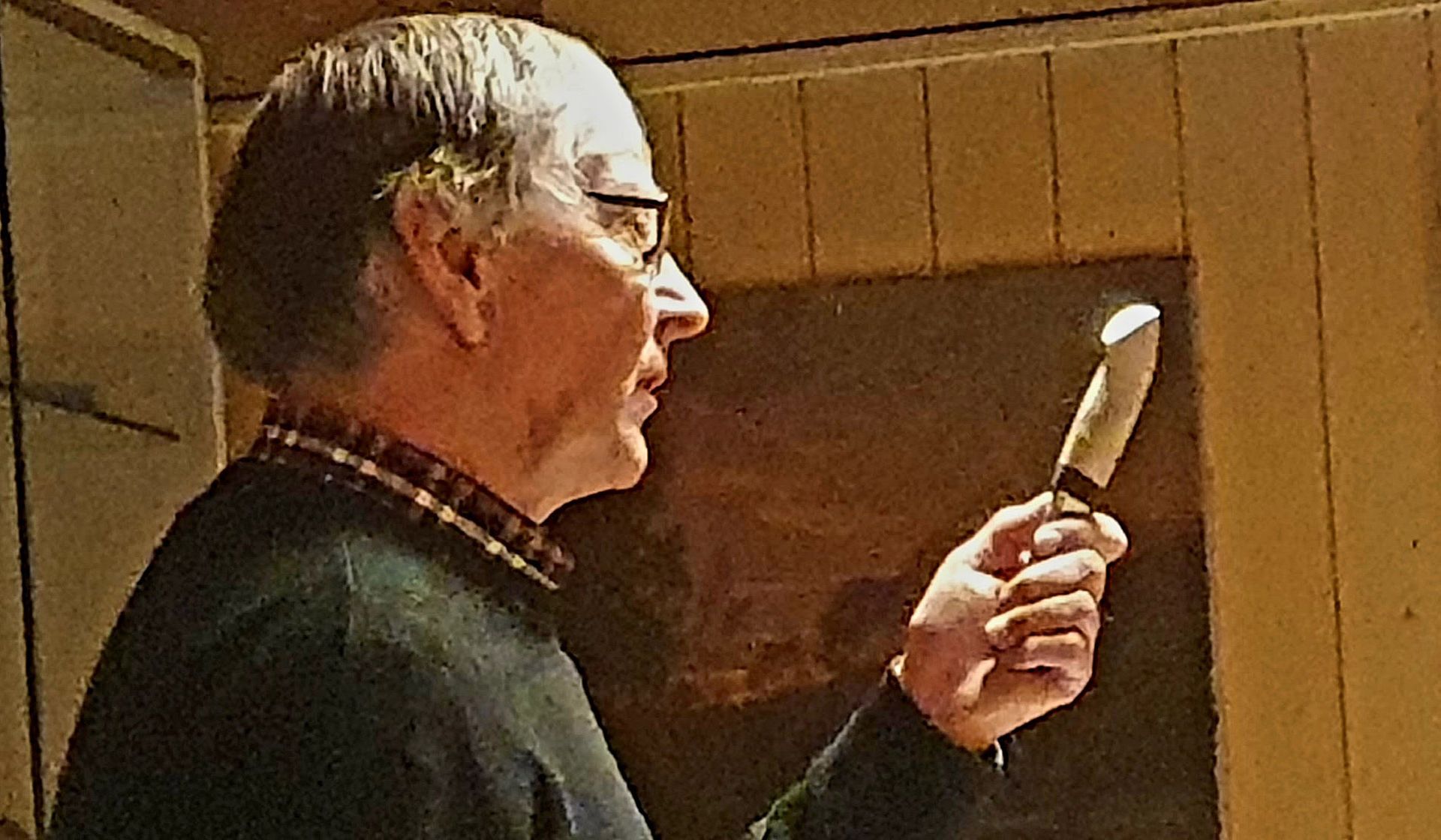 Mann med briller undersøker en kniv, innendørs, med trepanelvegg bak seg.