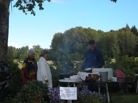 Folk ved en utendørs matbod med grill og bord, som serverer mat foran trær og en blå himmel.