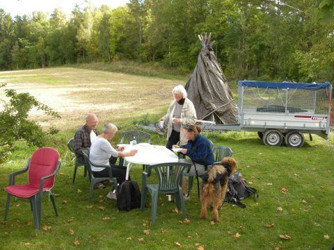 Folk sitter ved et bord og spiller et spill utendørs i nærheten av et tipi og en campingvogn, med en hund.