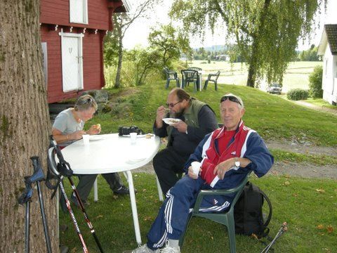 Tre personer sitter ved et bord og spiser utendørs. Rød bygning, gresskledd område og trær i bakgrunnen.