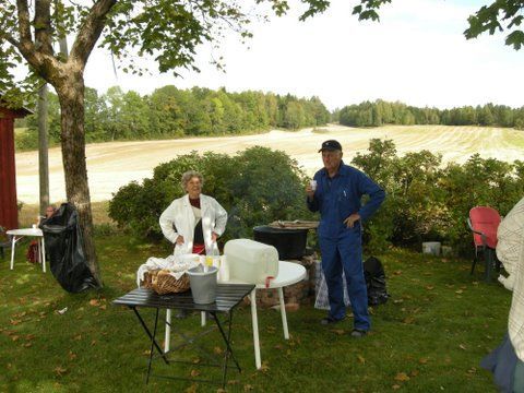 To personer, én i hvitt og én i blå kjeledress, står utendørs. Det er et bord med forsyninger og et jorde i bakgrunnen.