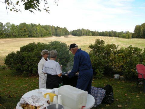 Tre personer griller utendørs, omgitt av busker og et jorde i det fjerne.
