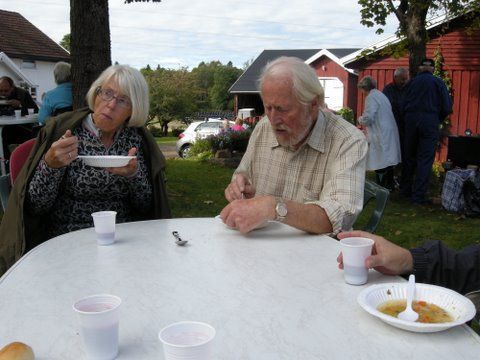 To eldre personer spiser suppe ved et utendørsbord, andre i bakgrunnen nær en rød bygning.