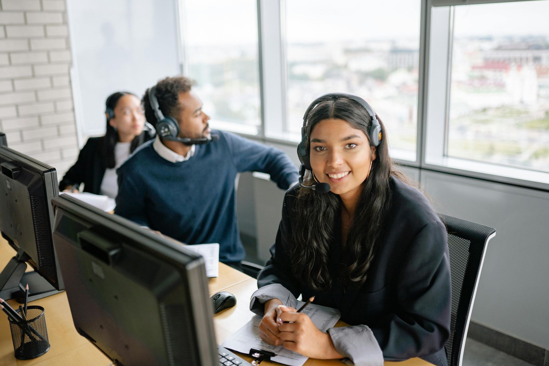 Three people in an office wearing headsets, smiling while working on computers.