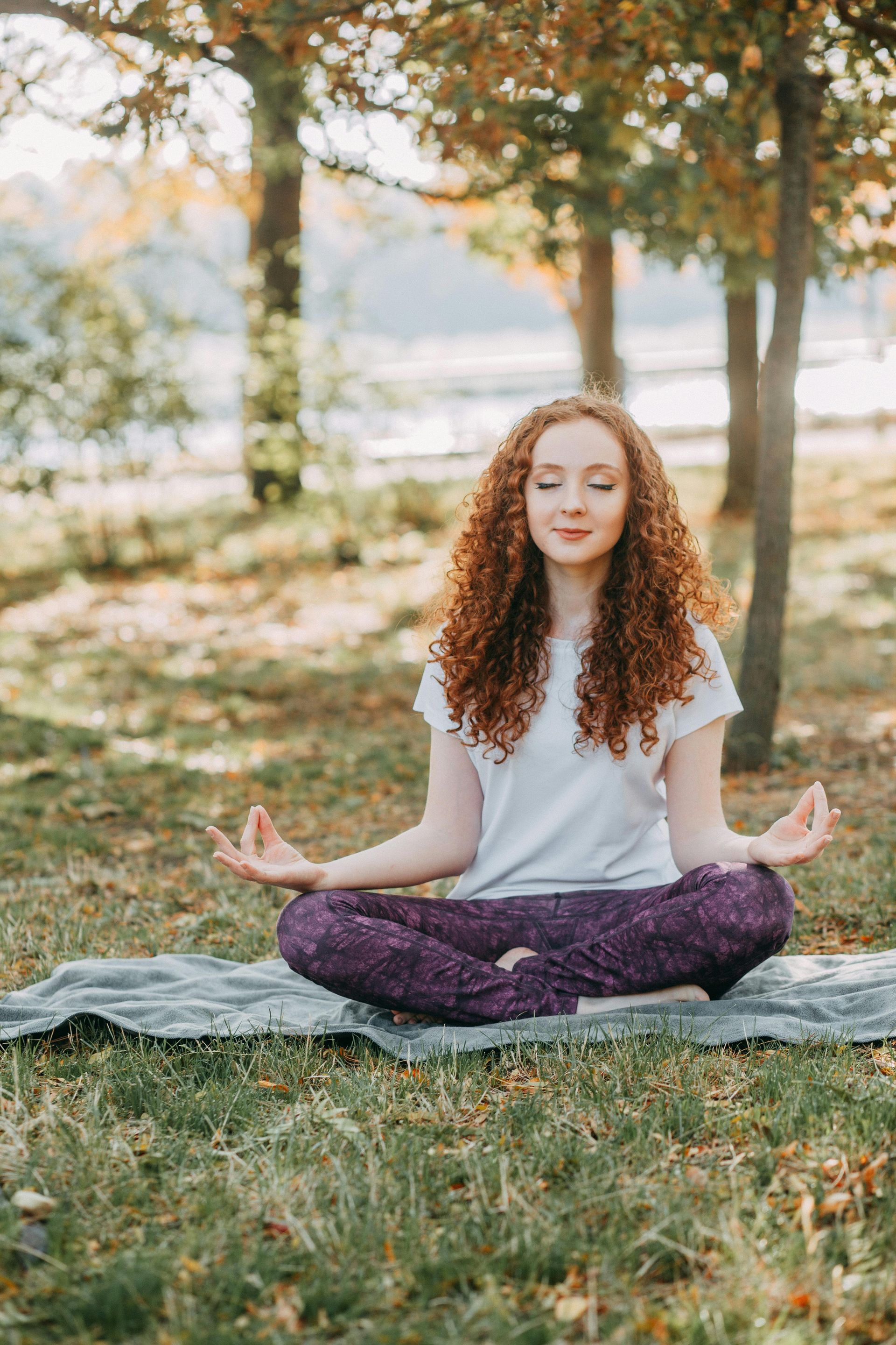 Woman with red curly hair meditating outdoors, eyes closed, hands in Gyan mudra. Wearing purple pants, light shirt.