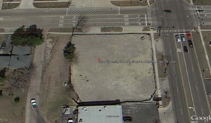 Aerial view of a vacant lot at an intersection with a building on the bottom, trees, and roads.
