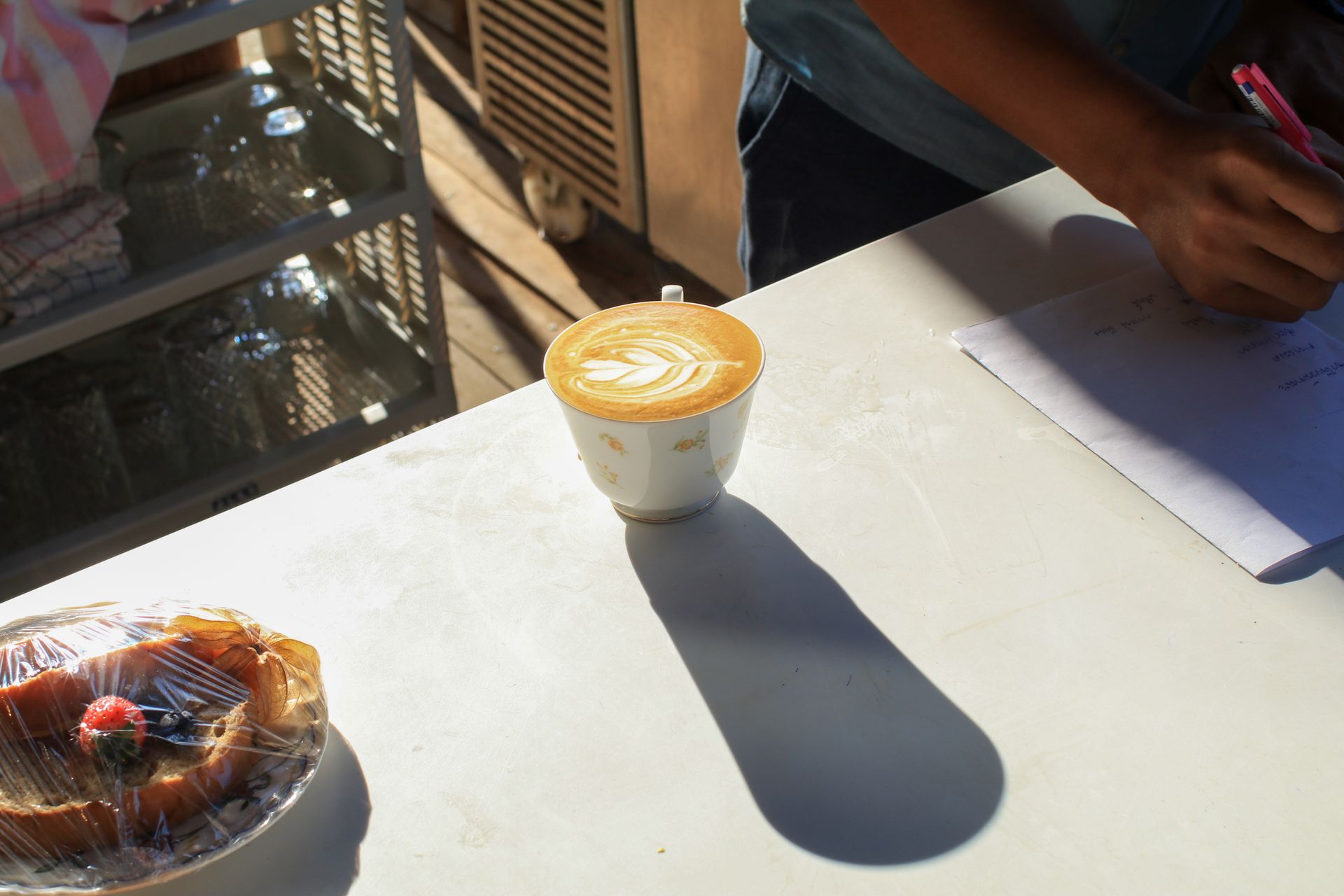 Coffee cup and pastry on a sunlit white table, with a notebook and person’s hands nearby.