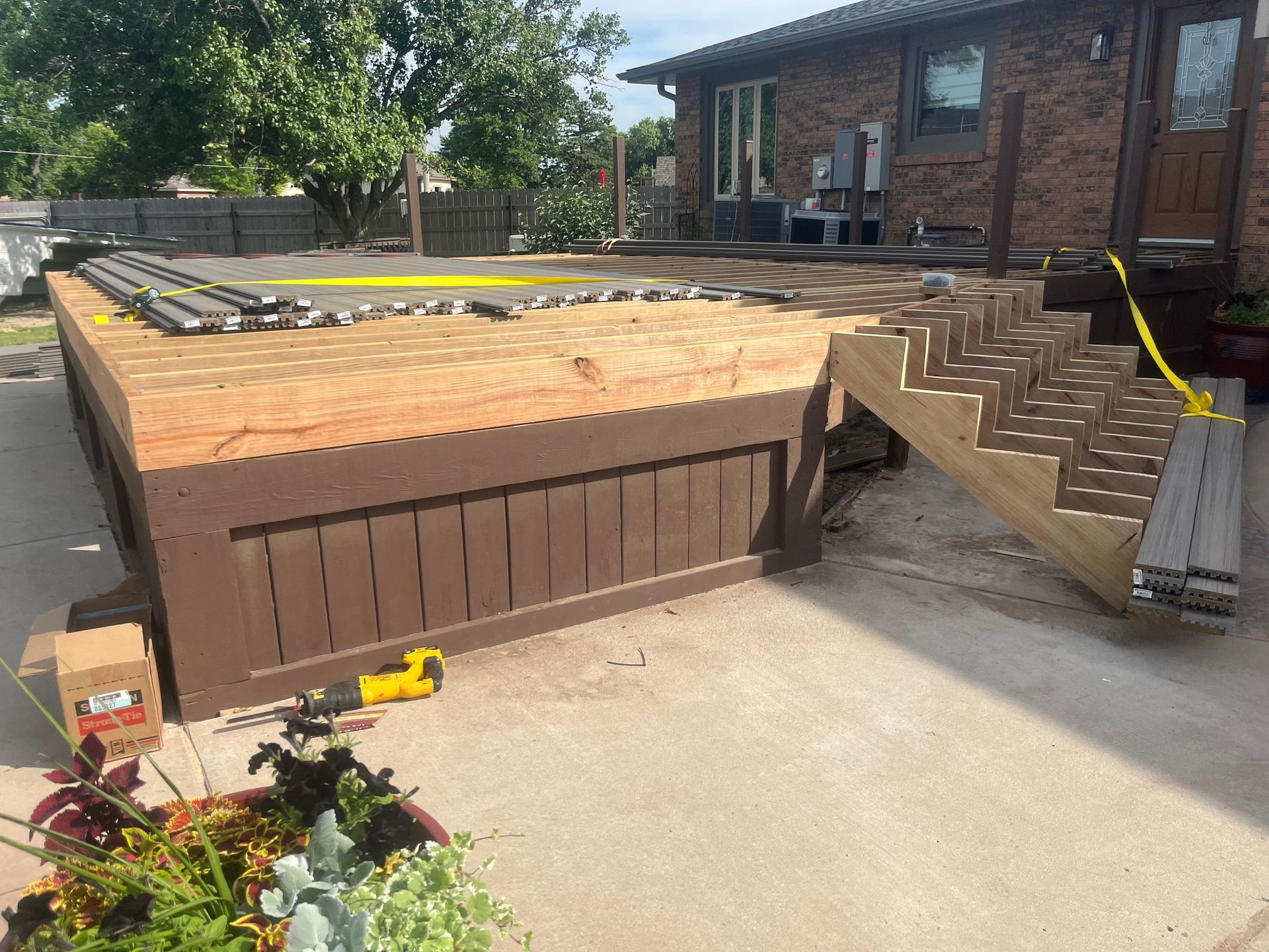 An unfinished wooden deck under construction with exposed plywood, a brown-painted base, and a set of stairs on a patio.