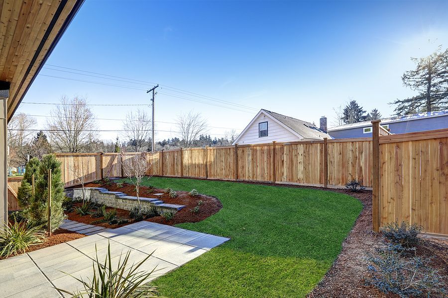 A backyard scene with a small concrete patio, a patch of green lawn, a terraced planter bed, and a tall wood fence.