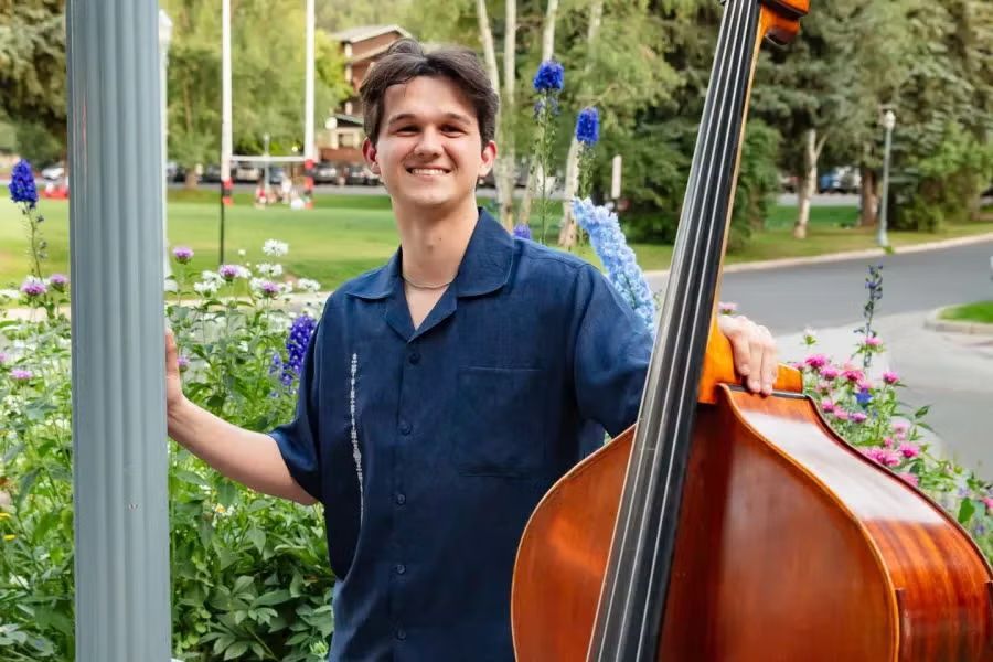 A smiling person in a blue shirt standing outdoors next to a large upright bass with a park and trees in the background.
