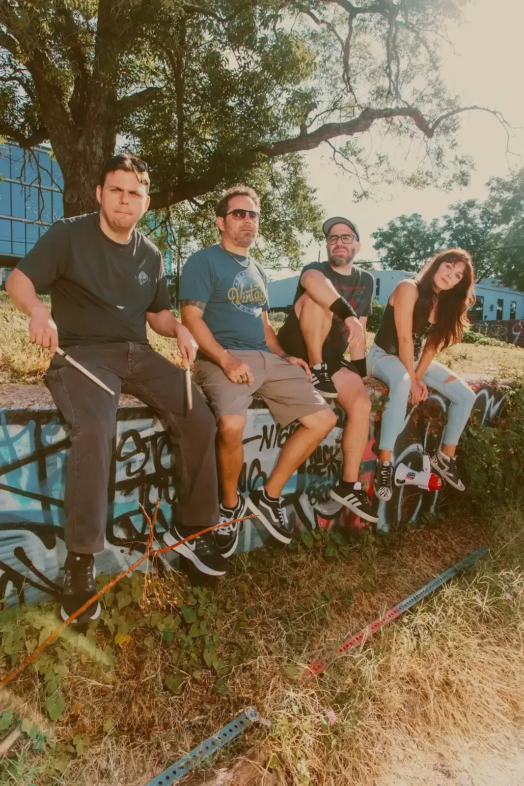 Four people sit on a graffiti-covered wall under a tree on a sunny day. They are wearing casual clothing.
