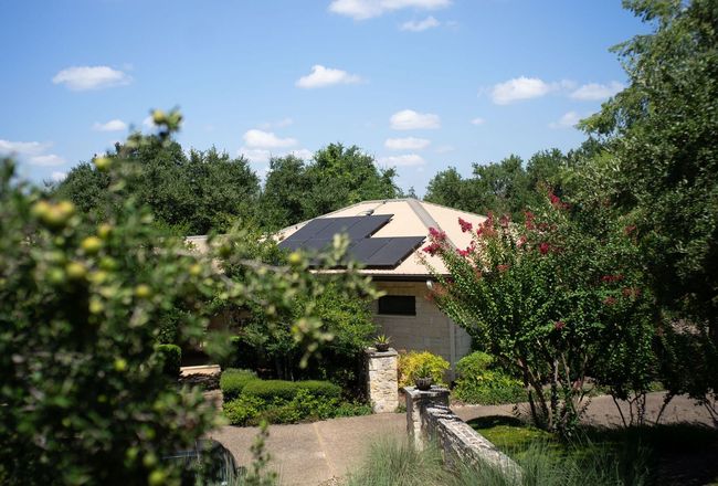 A house with solar panels on the roof is surrounded by trees and bushes.