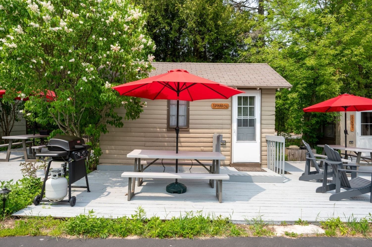 Cabin with red umbrellas, picnic table, grill, and chairs on a wooden deck.