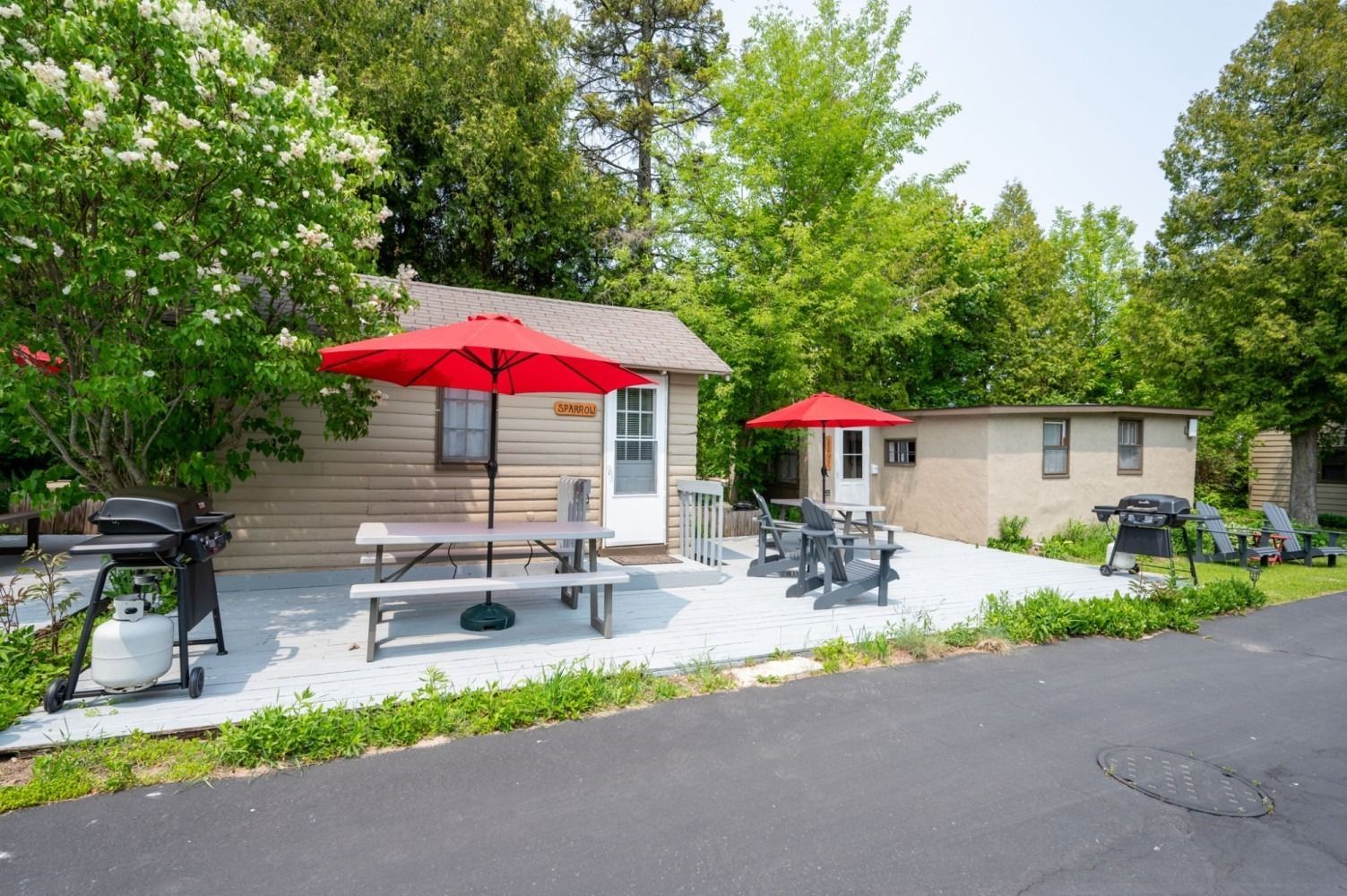 Small cabin with red umbrellas, picnic table, and grills on a paved patio. Trees in background.