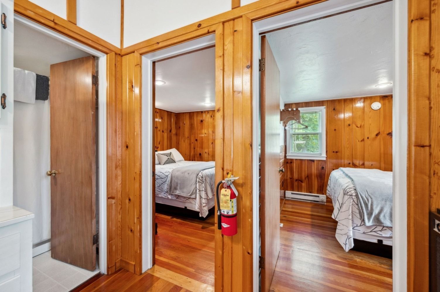 Hallway with three doorways, two to bedrooms with beds, one to a bathroom with a white cabinet.