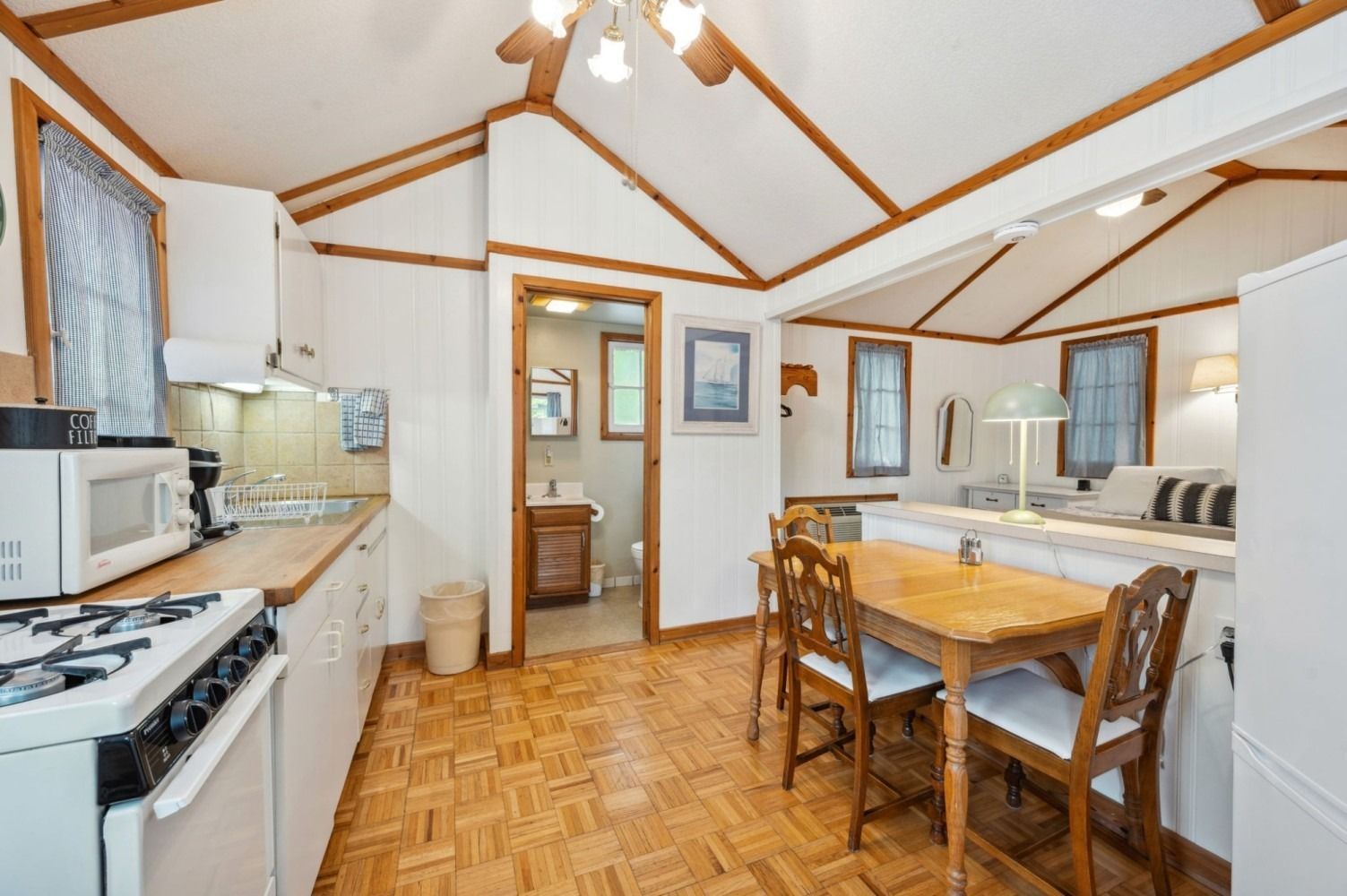 Small kitchen with wooden floors, dining table, and view to bathroom. Bright with white walls and natural light.