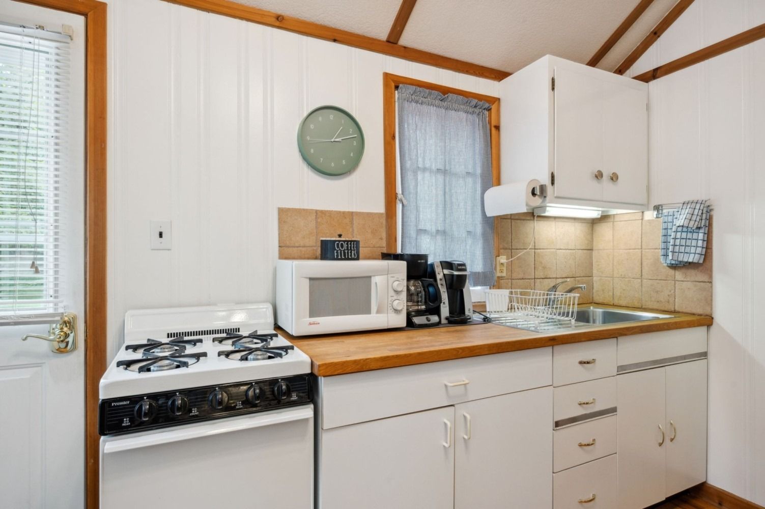 Small kitchen with white cabinets, stove, microwave, and sink; a window and a clock are also visible.