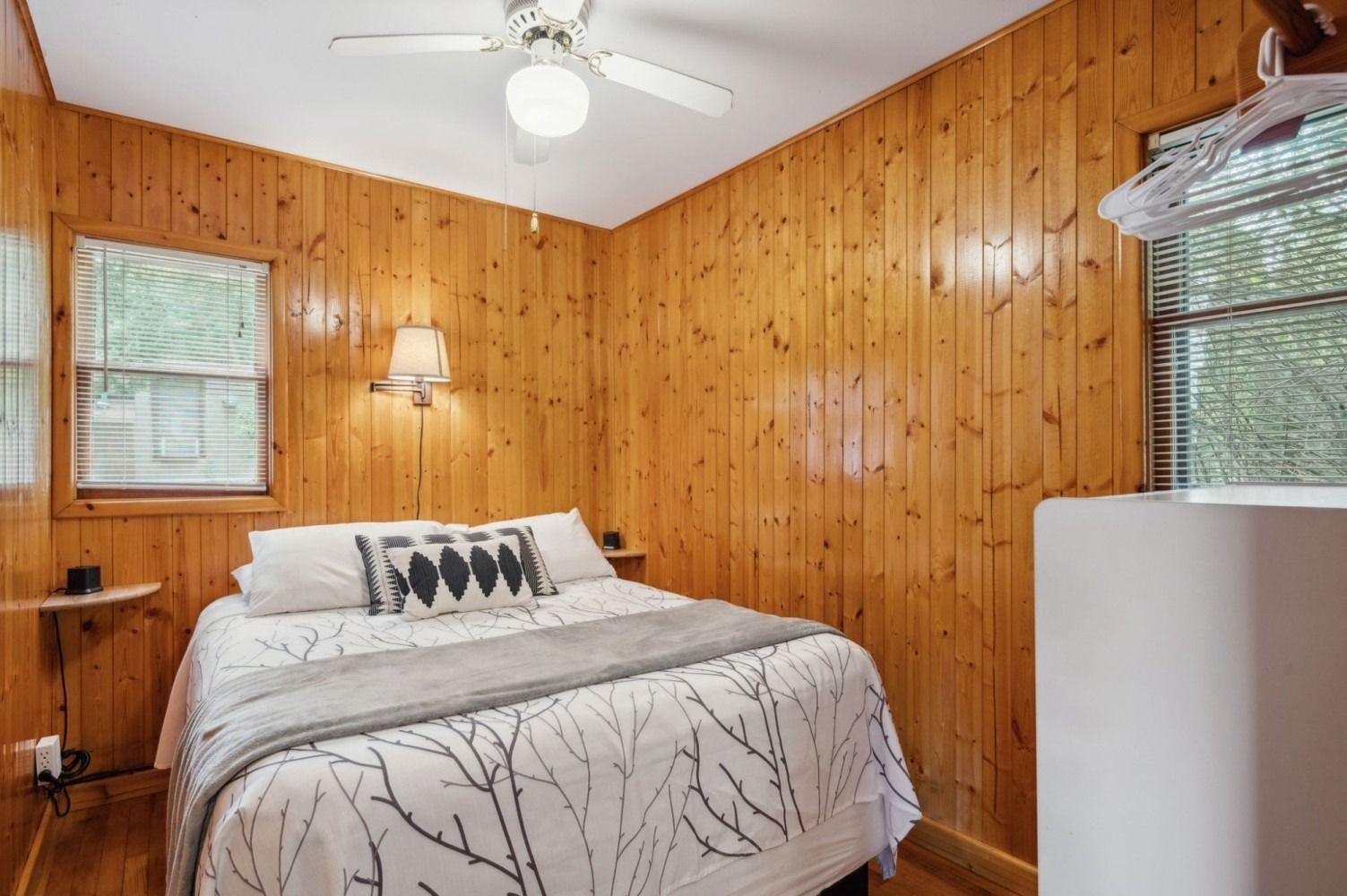 Bedroom with wood-paneled walls, bed with gray and white bedding, window with blinds, ceiling fan.