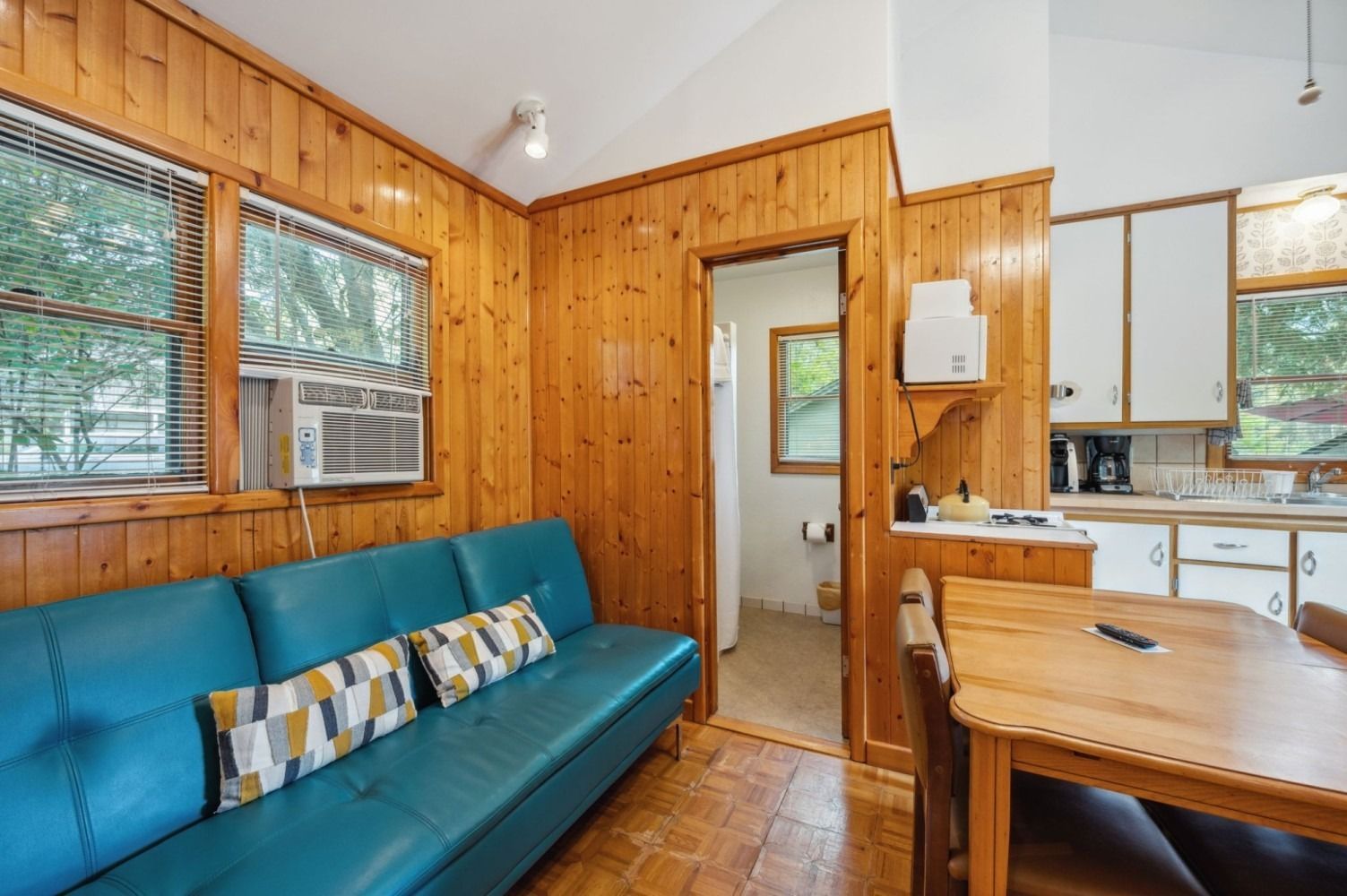 Cozy cabin interior with turquoise couch, wood paneling, small kitchen, and a glimpse into the bathroom.