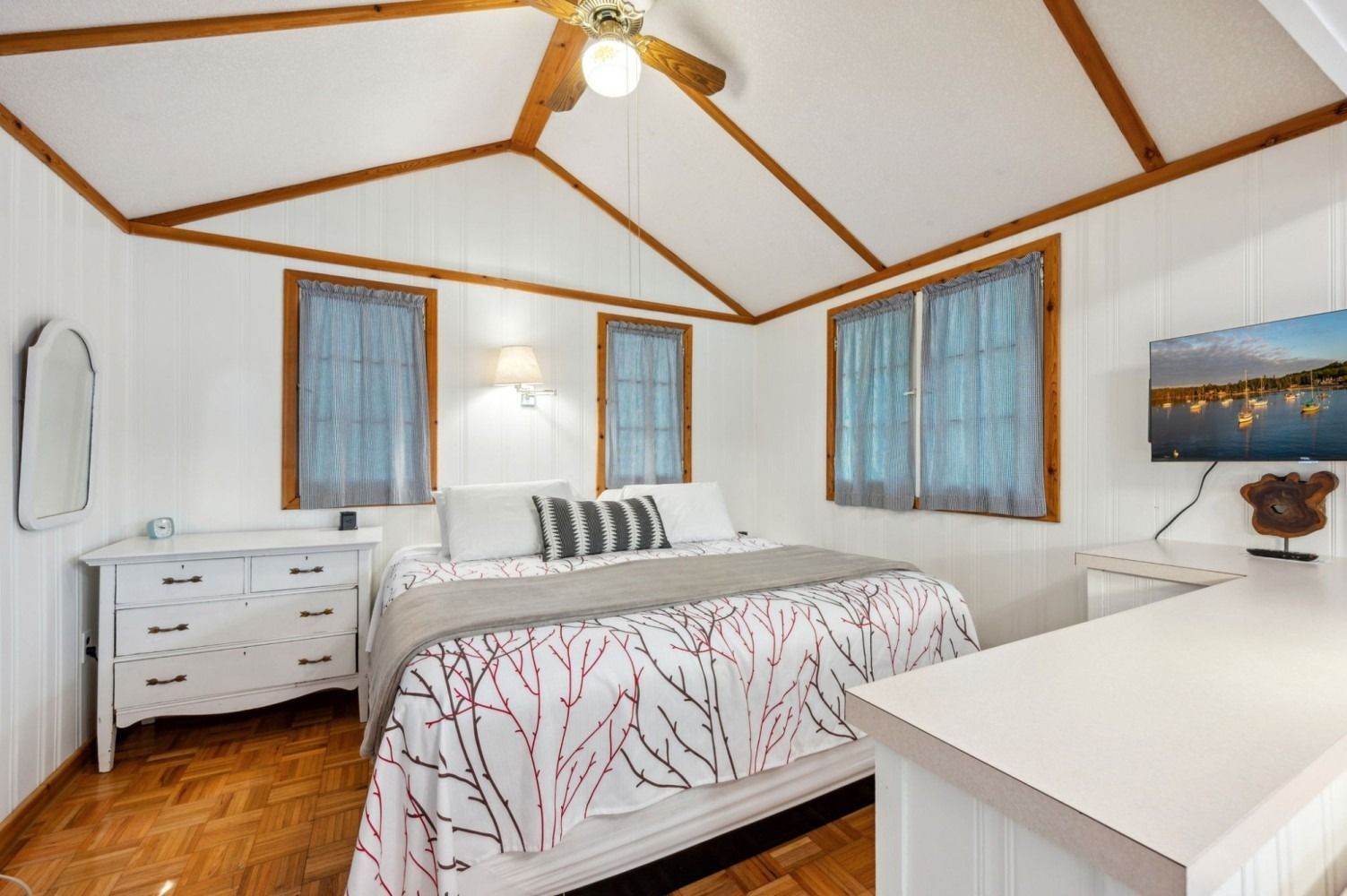 Bedroom with a bed, white walls, wood trim, windows with blue curtains, and a TV.