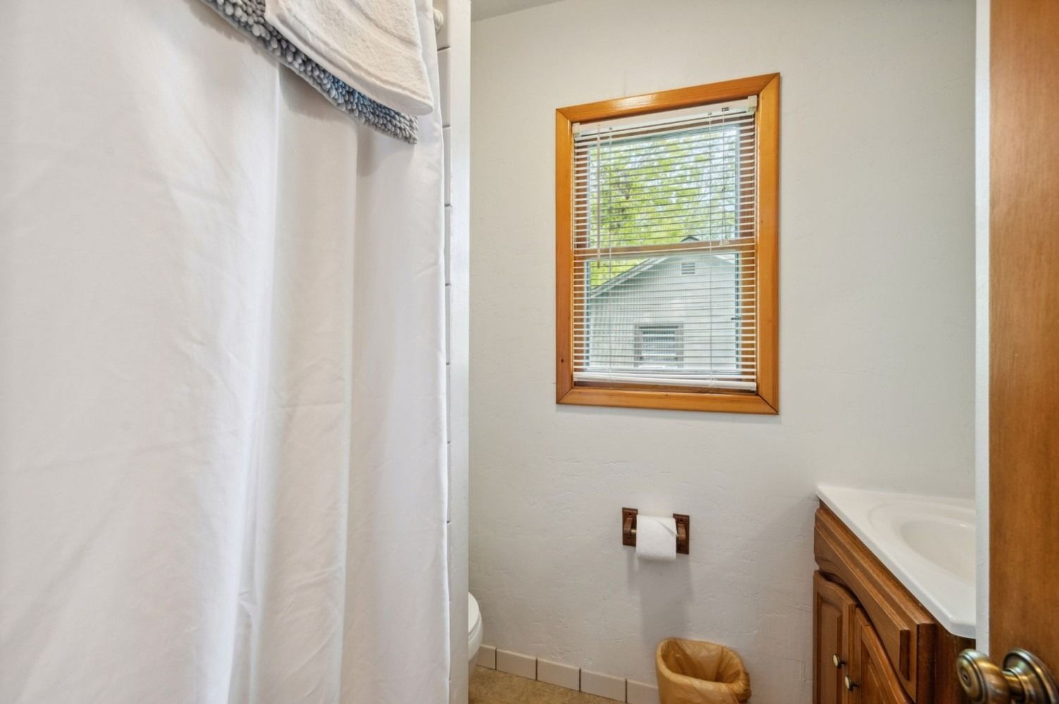 Bathroom interior with white shower curtain, window with blinds, brown vanity, and a trash can.