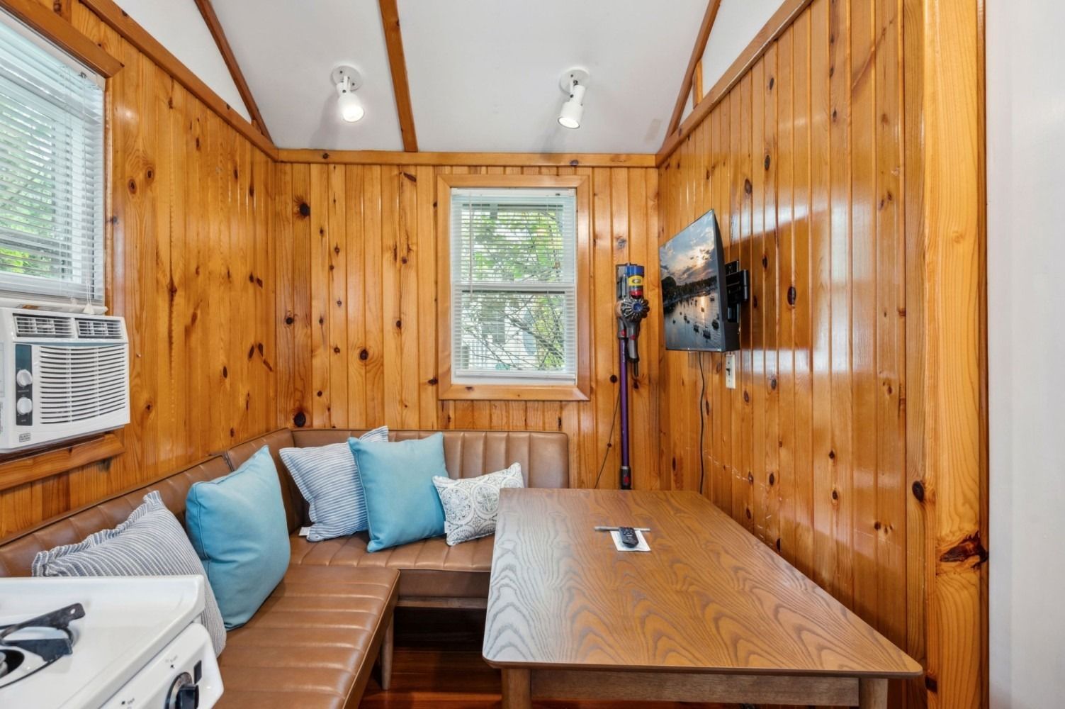 Small cabin dining area with wood paneling, L-shaped bench seating, table, and mounted TV.