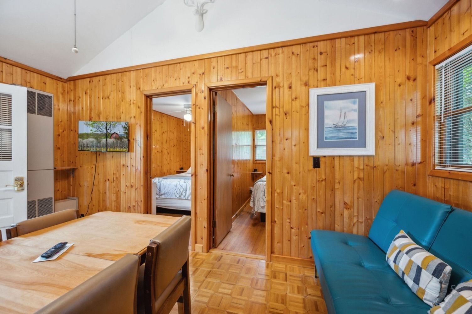 Interior of a cabin with wood paneling, featuring a dining table, teal sofa, and doorways to bedrooms.