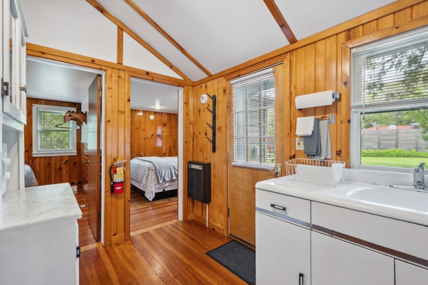 Interior view of a small cabin kitchen with wooden walls, a white sink, and a view into the bedroom.