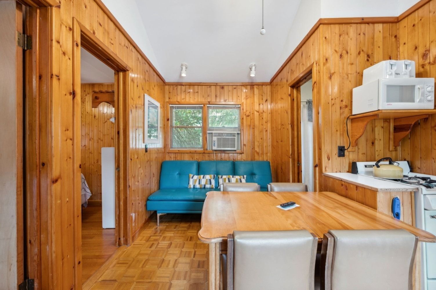 Cozy cabin interior: wood-paneled walls, blue couch, small kitchen with microwave, table, chairs, doorway to a bedroom.