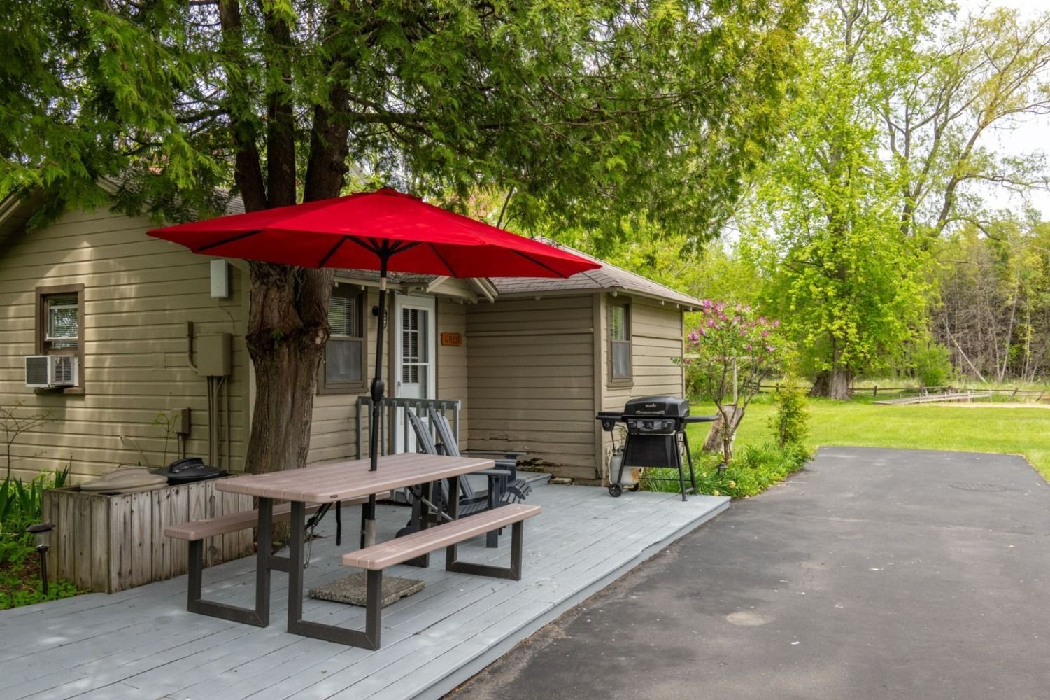 Cottage with red umbrella over picnic table on deck, next to a paved driveway.