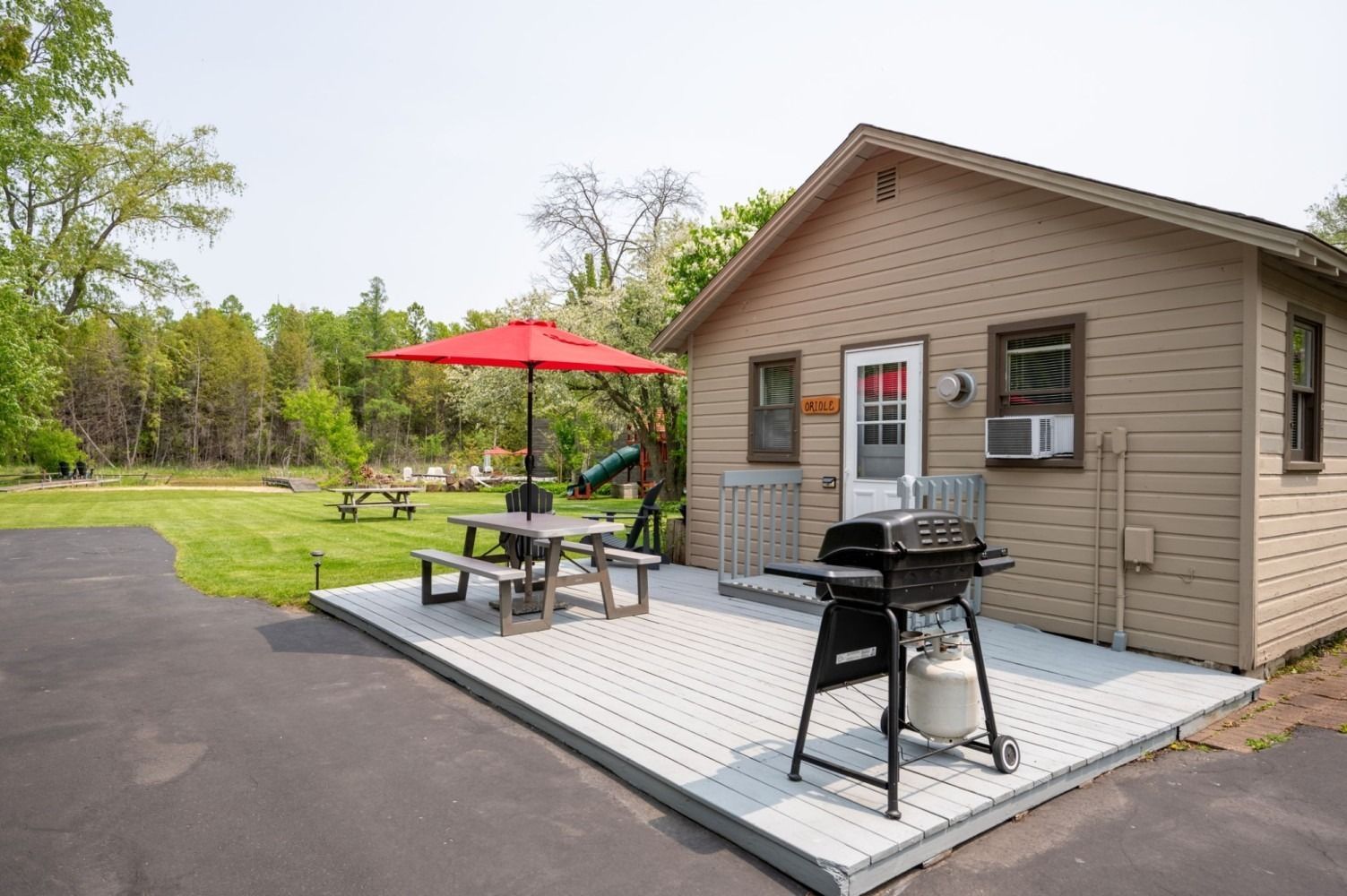 Tan cabin with a gray deck, picnic table, red umbrella, and grill. Lawn and trees in background.