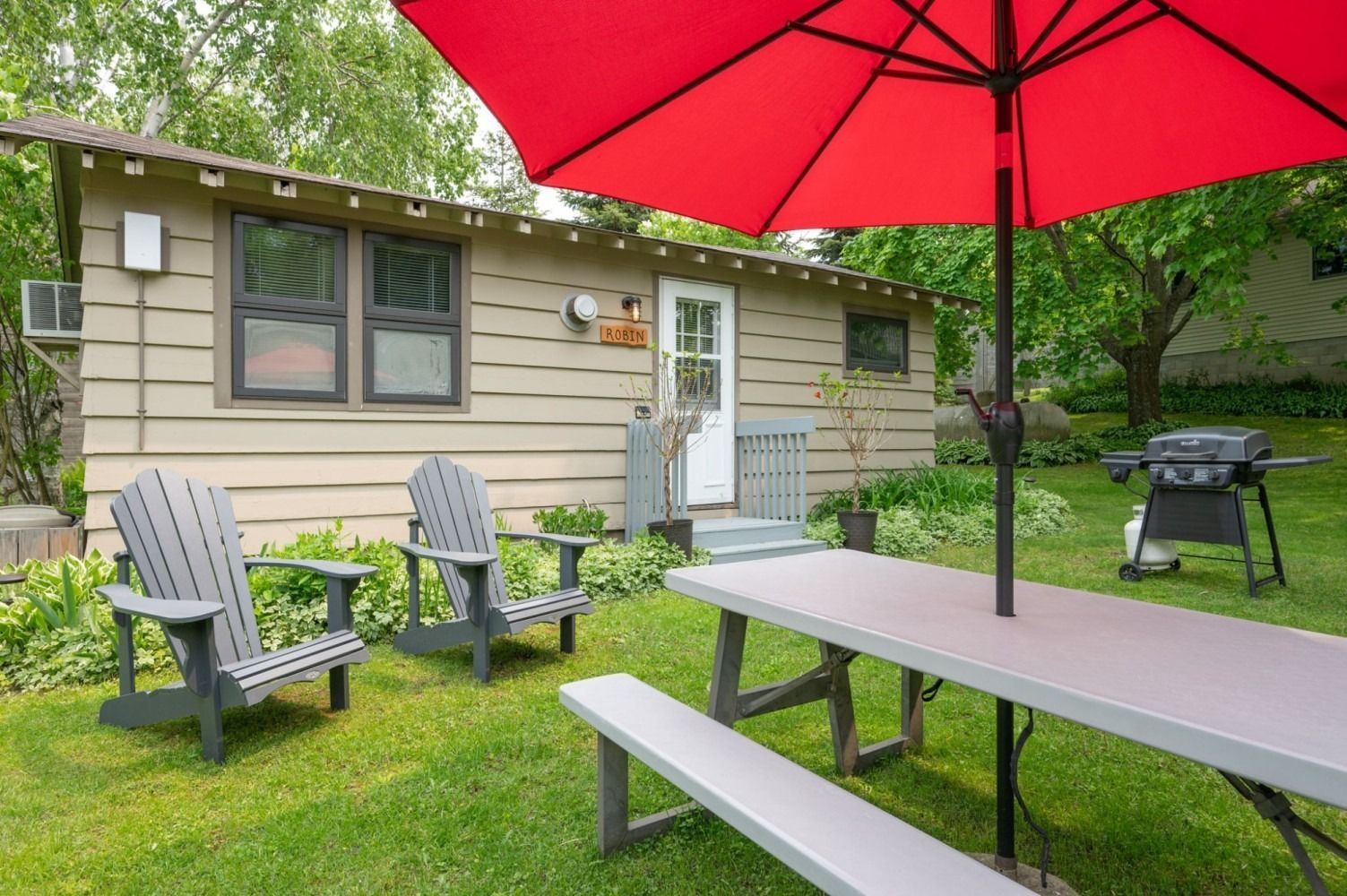 A small cabin with outdoor seating under a red umbrella and a grill in a grassy yard.