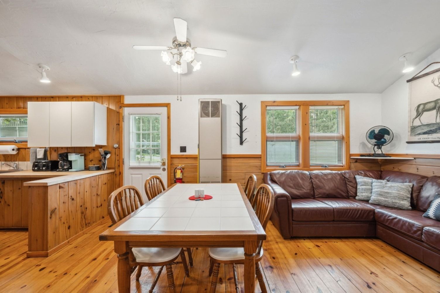 Open-plan living space with kitchen, dining area, and leather sectional. Pine wood paneling, windows, and ceiling fan.