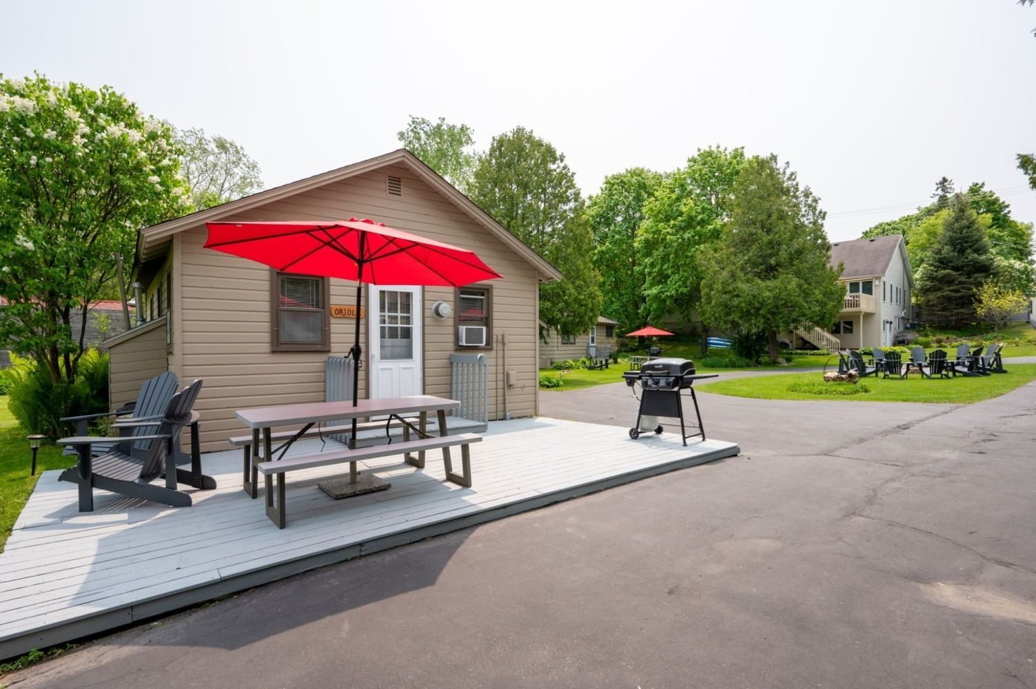 Cottage with a red umbrella, picnic table, and grill on a deck. Trees and a path are nearby.