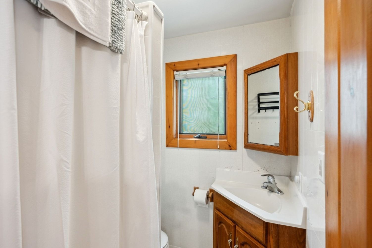 Small bathroom with white walls, vanity, mirror, shower curtain, and window with wooden trim.