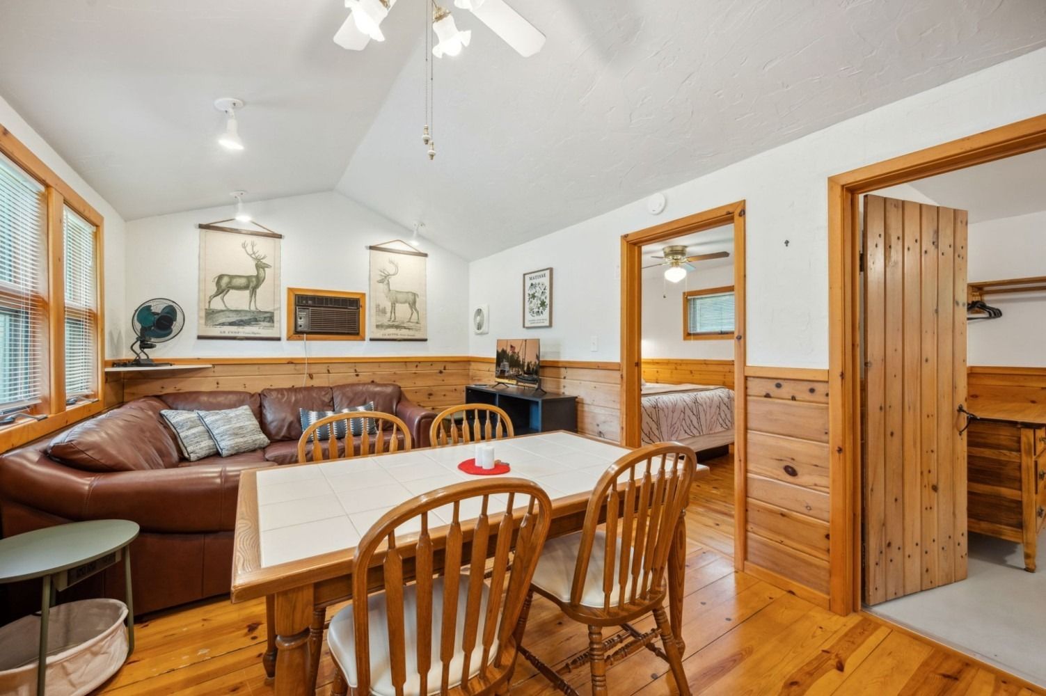Cozy cabin interior: dining table, brown sofa, wood paneling, open door to bedroom.