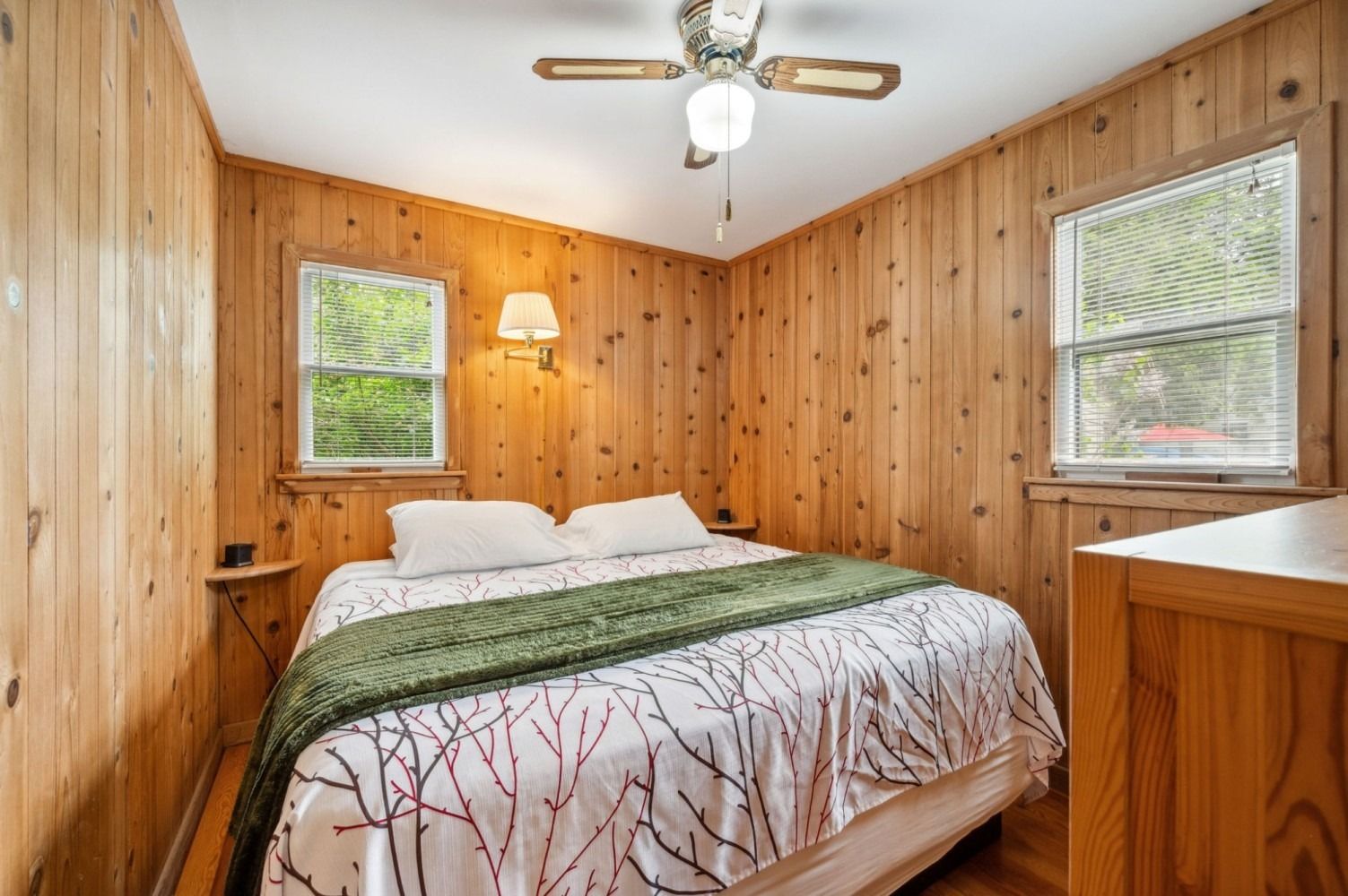 Bedroom with wood-paneled walls, bed with green blanket, two windows, and a ceiling fan.