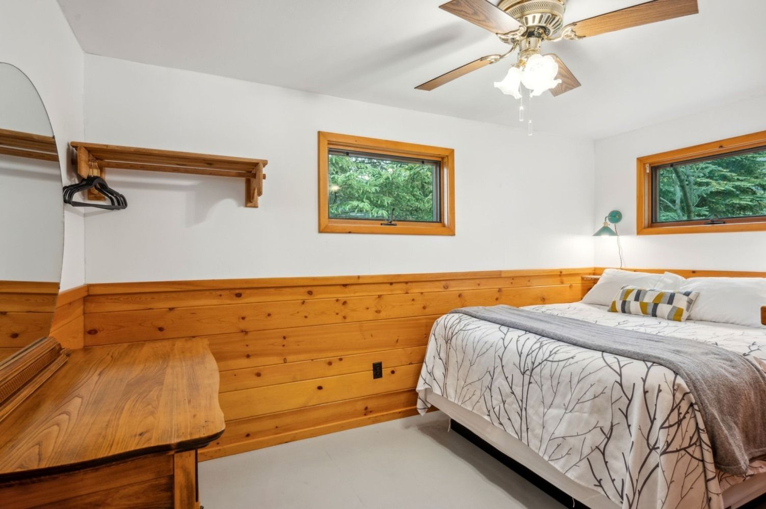 Bedroom with wooden paneling, white walls, bed with gray blanket, two windows, and dresser with mirror.