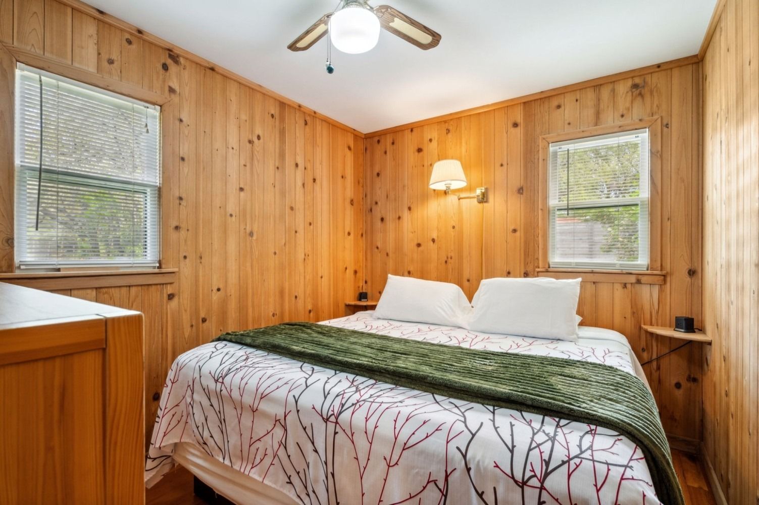 Bedroom with wood-paneled walls, a bed with a white and red patterned comforter, and two windows.