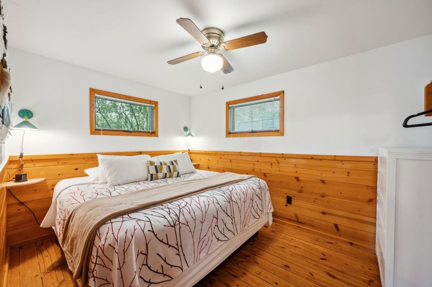 Bedroom with wood paneling, a bed, two windows, and a ceiling fan.
