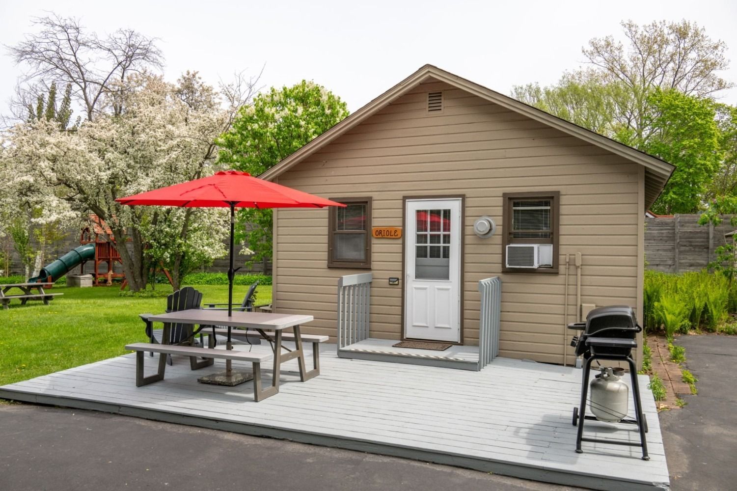 Small tan cottage with a red umbrella over a picnic table on a gray patio. Grill on the right.